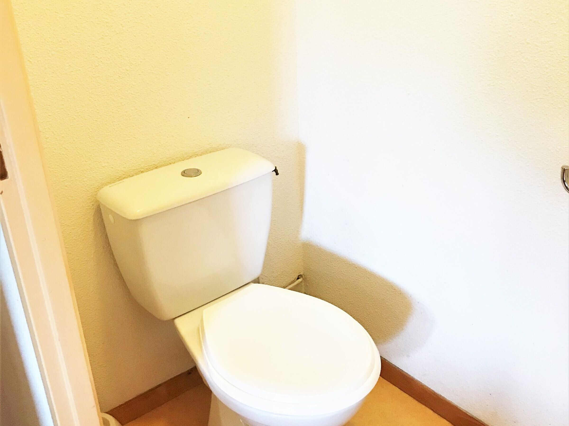 A white toilet in a corner of a room with a plain wall and a wooden floor.