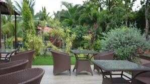 An outdoor patio area with wicker furniture and a glass table, surrounded by lush greenery and a red-roofed building in the background. - The Grand Neelam Hotel (Raipur)