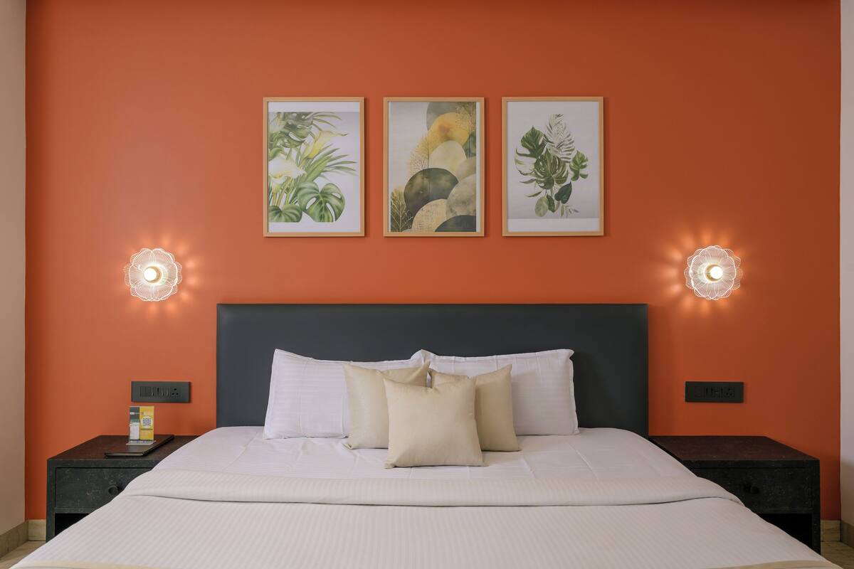 A neatly made bed with white linens and three pillows, a dark headboard, and framed botanical artwork on the orange wall above.