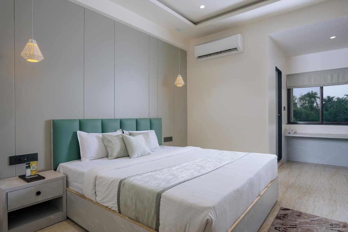A modern bedroom with a large bed, a nightstand, a wall-mounted air conditioner, and a window with a view of greenery.