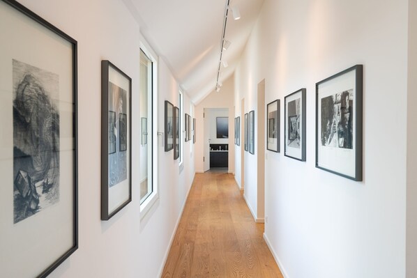 A hallway with framed artworks on the walls, wooden flooring, and a kitchen area in the background.