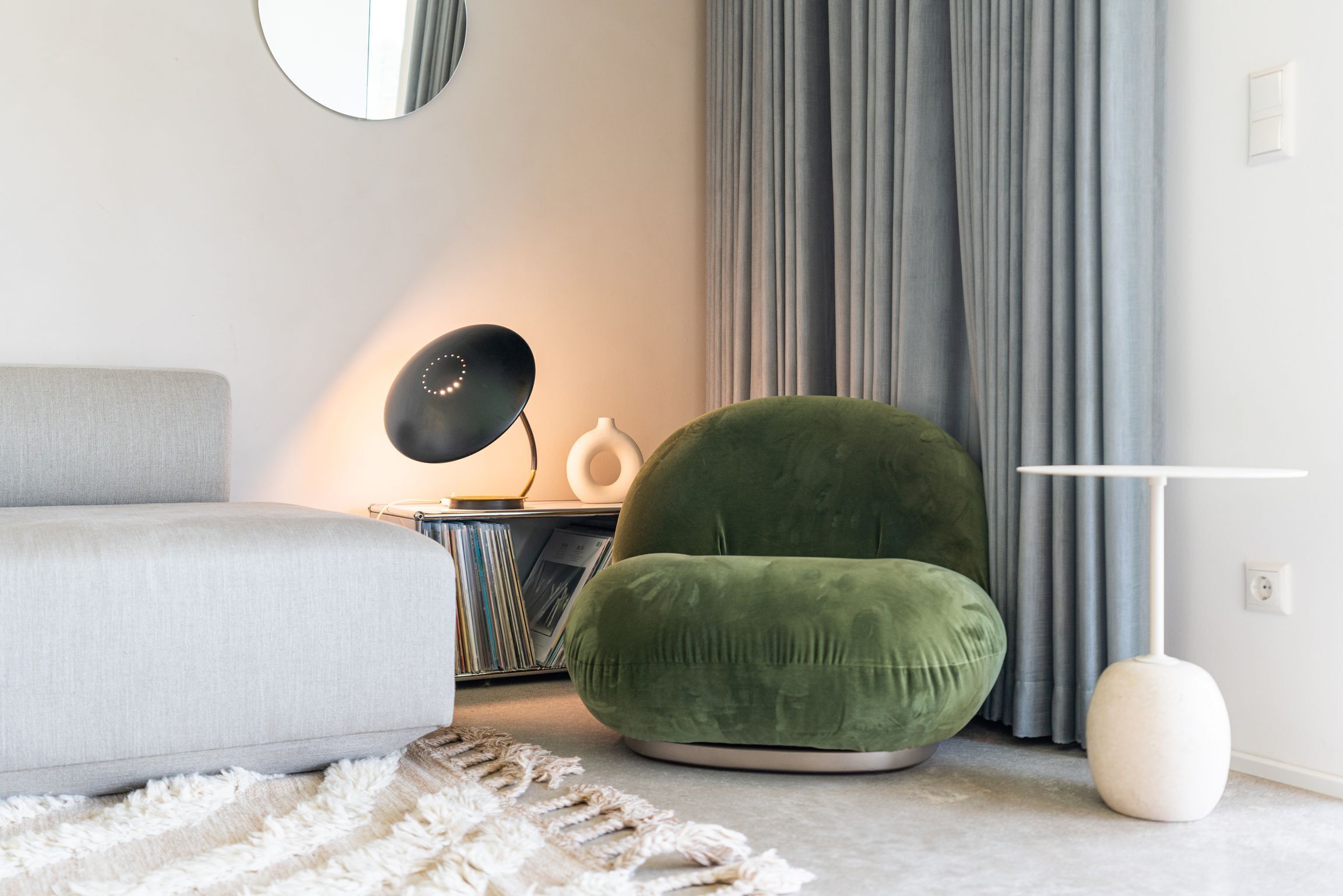 Un salon moderne avec un canapé gris, un fauteuil vert, une petite table d’appoint avec une lampe et un lampadaire blanc.