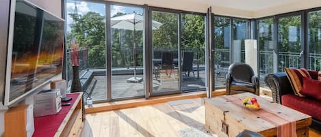 A spacious living room with a large TV, wooden flooring, a red sofa, a coffee table, and a view of the outdoors through large glass doors.