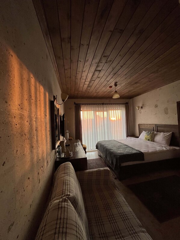 A room with wooden ceiling beams, a bed, a sofa, a desk with a lamp, and a window with curtains. - UCHI Cappadocia (Nevsehir)