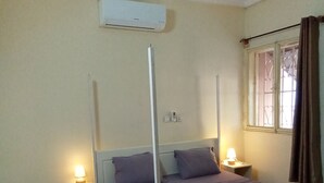 A bedroom with a bed, two bedside tables with lamps, an air conditioning unit, a window with curtains, and a ceiling fan.