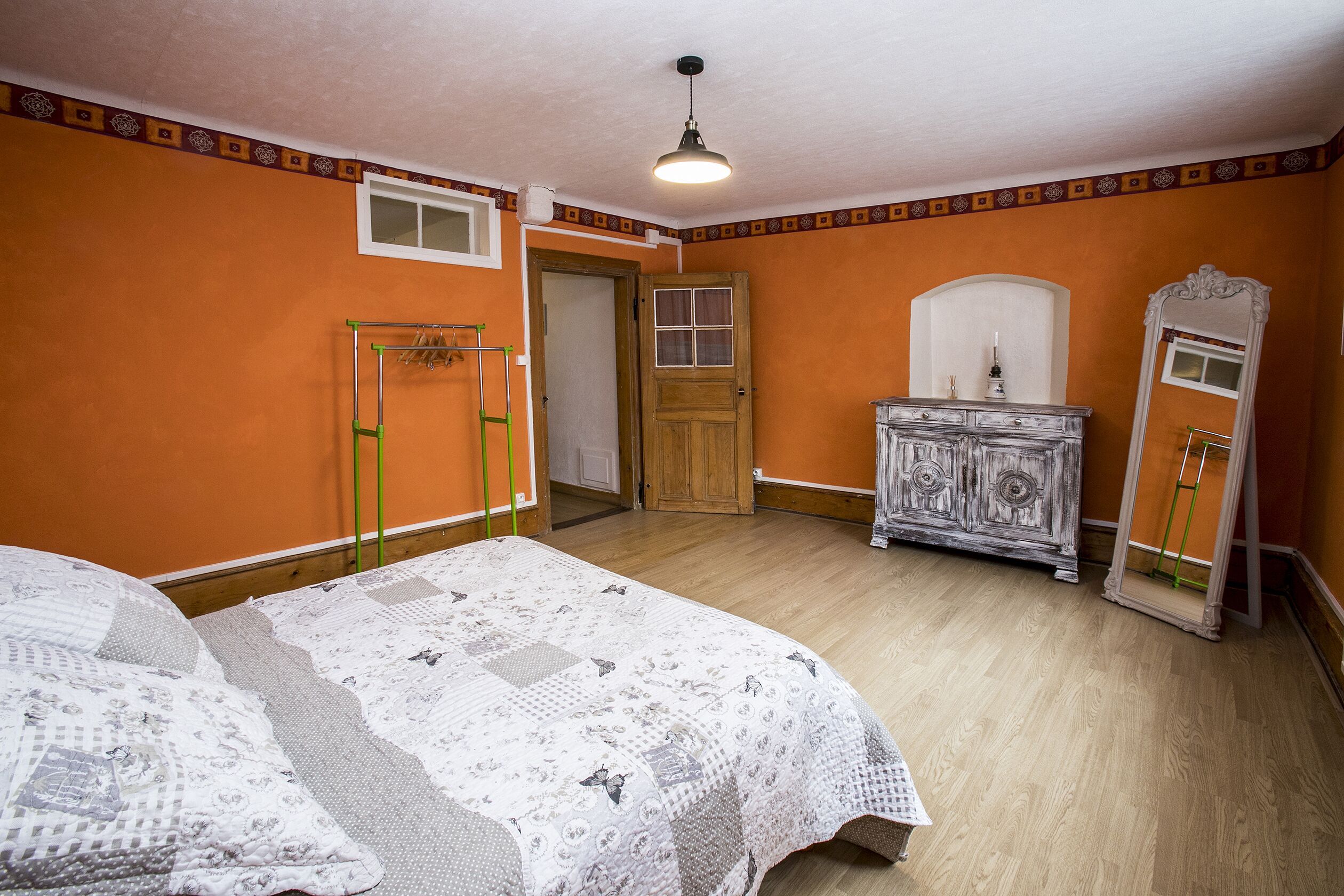 Une chambre avec un mur orange, une porte en bois, une commode, un miroir et un lit recouvert d’une couvre-lit à motifs.