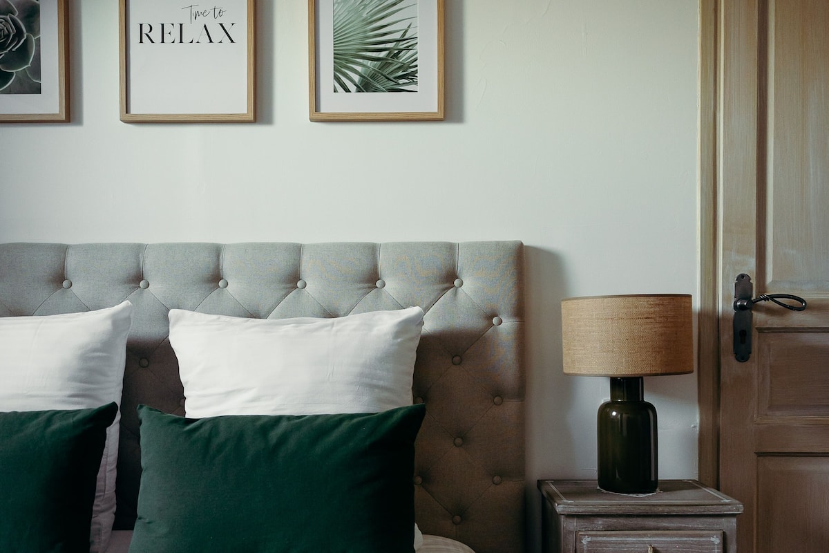 Une chambre à coucher avec une tête de lit capitonnée, une table de chevet avec une lampe et des œuvres d’art encadrées accrochées au mur.
