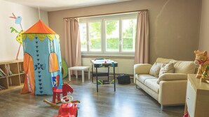 Una sala de estar con una carpa infantil, un caballo balancín, una mesa con un juego de mesa, un sofá y una ventana con cortinas.