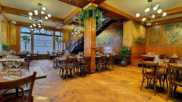 A restaurant with wooden floors, multiple dining tables, and chairs. There is a staircase with a railing and a large plant in the center. - Le Tonnelier (Bulle)