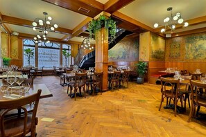 A restaurant with wooden floors, multiple dining tables, and chairs. There is a staircase with a railing and a large plant in the center.