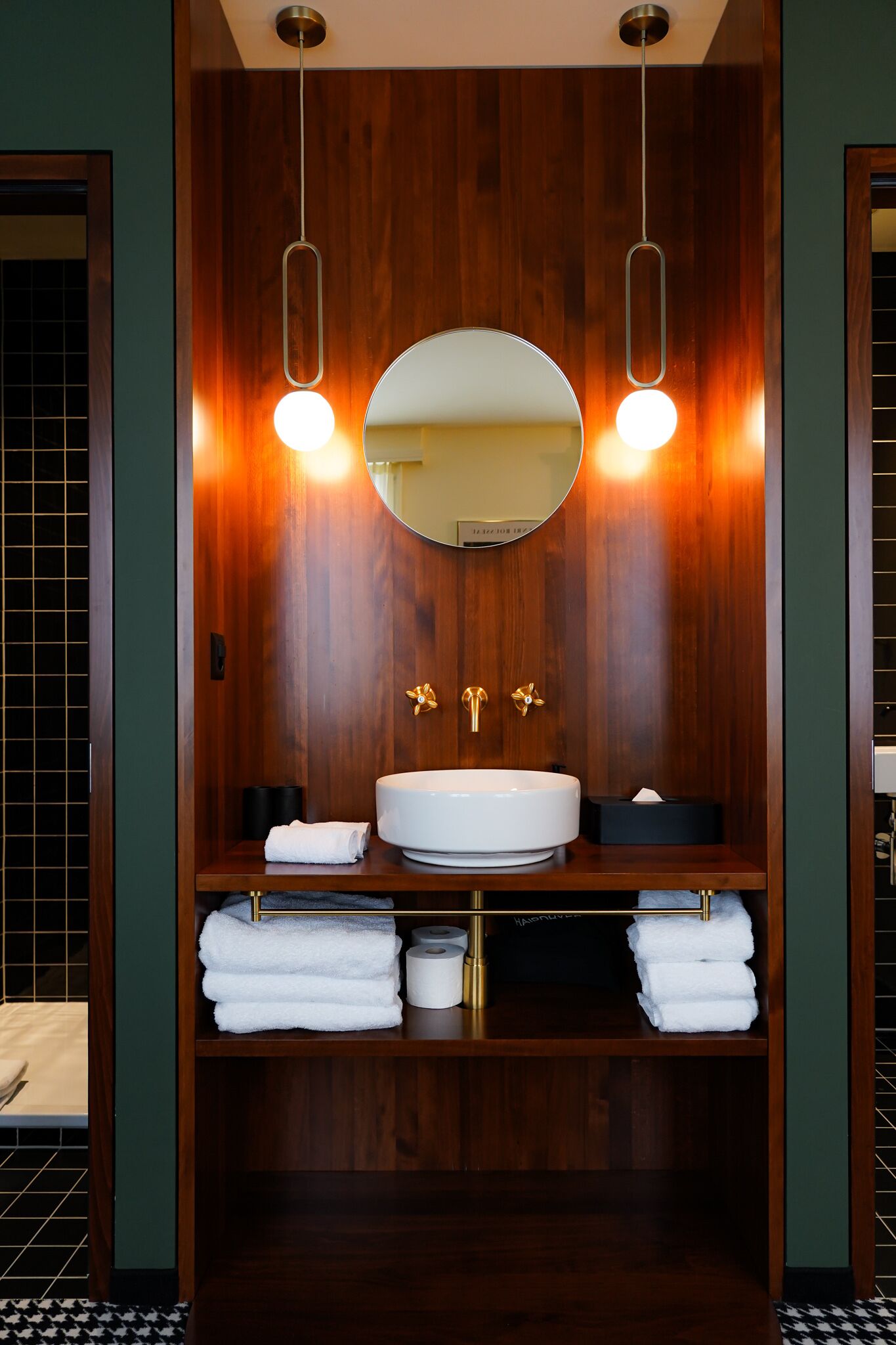 Un baño con tocador de madera, un espejo redondo, dos luces empotradas en la pared y un lavabo con grifo dorado.