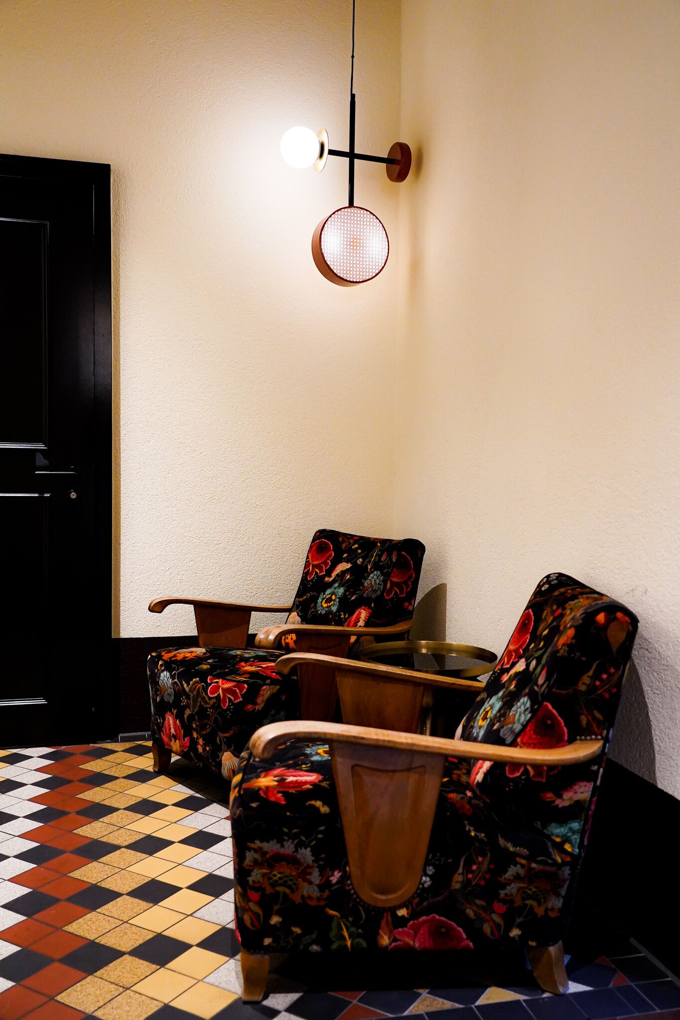 Una habitación con piso a cuadros, dos sillones con tapicería floral, una mesa redonda y una lámpara fijada en la pared.