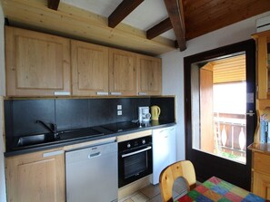 Uma cozinha compacta com armários de madeira, bancada preta, lava-louças, forno e uma janela com vista para uma varanda.