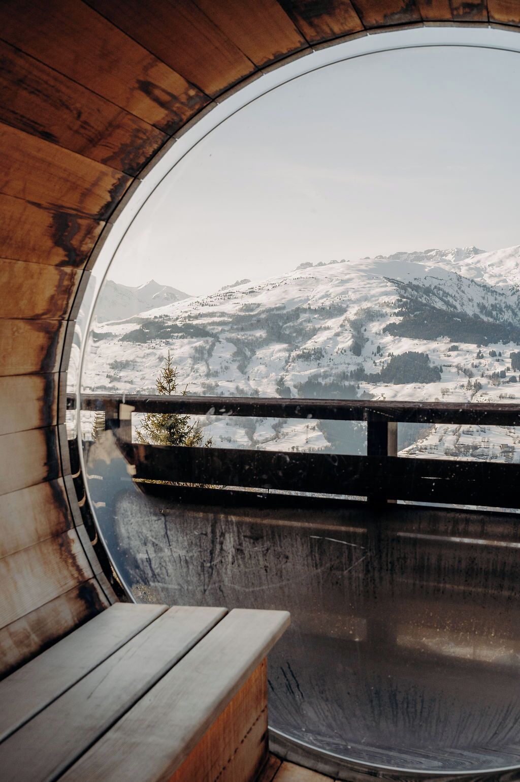 Une fenêtre ronde offrant une vue sur une montagne enneigée, un banc en bois et une rambarde en métal.