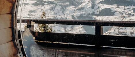 Une fenêtre ronde offrant une vue sur une montagne enneigée, un banc en bois et une rambarde en métal.