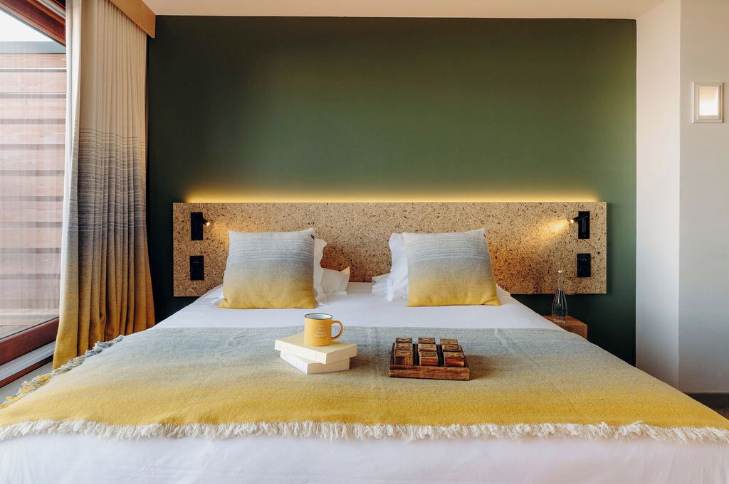 Habitación de hotel con cama, cabecero con luz, mesita de noche, ventana con cortinas y vistas al exterior.