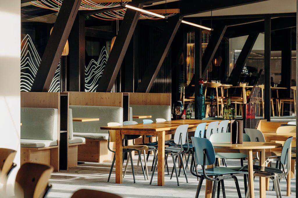 Un espace de restauration moderne avec des tables et des chaises en bois, un plafond haut avec des poutres apparentes et une vue sur la ville grâce à de grandes fenêtres.
