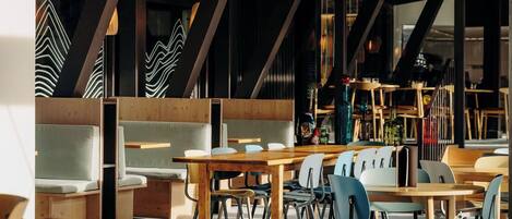 Un espace de restauration moderne avec des tables et des chaises en bois, un plafond haut avec des poutres apparentes et une vue sur la ville grâce à de grandes fenêtres.