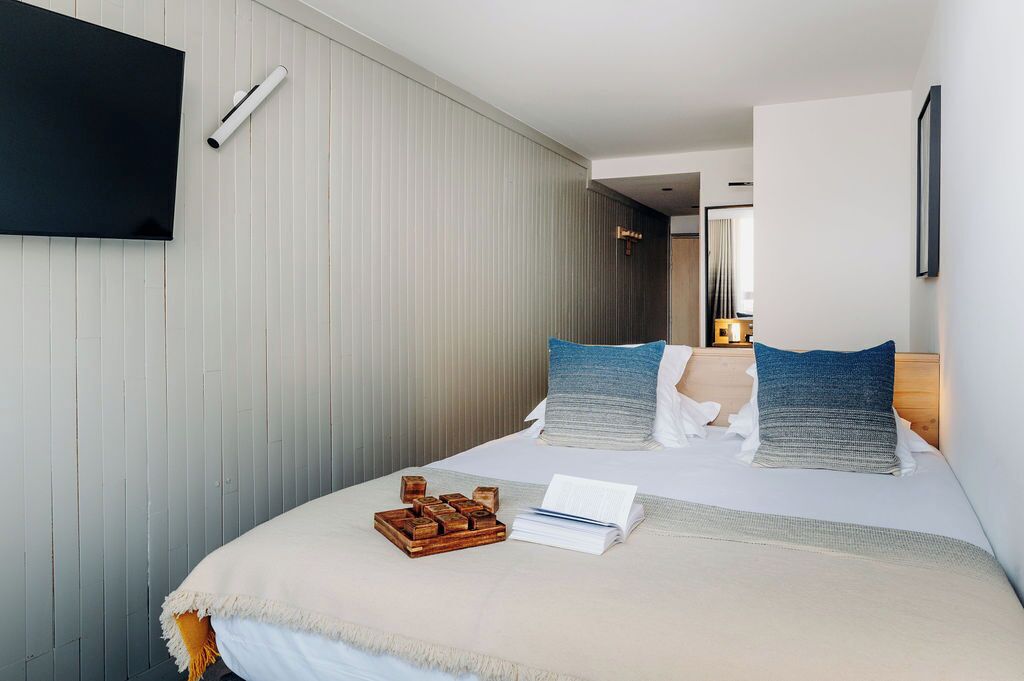 Una habitación de hotel moderna con una cama grande, un televisor de pantalla plana montado en la pared y una mesita de madera con un libro y una bandeja de bloques de madera.