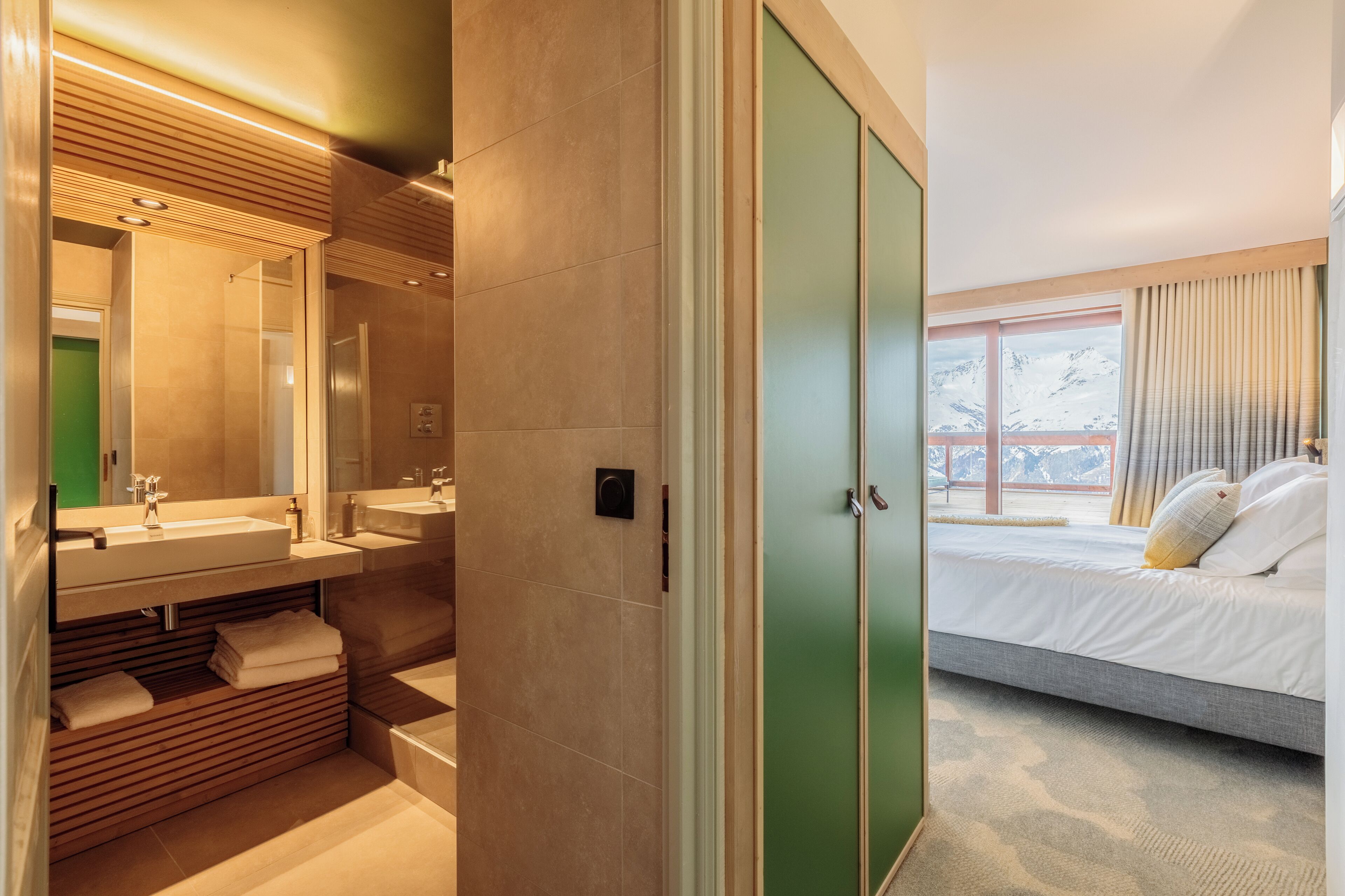 Una habitación de hotel moderna con una cama grande, un baño independiente con tocador y vistas a montañas nevadas a través del ventanal.