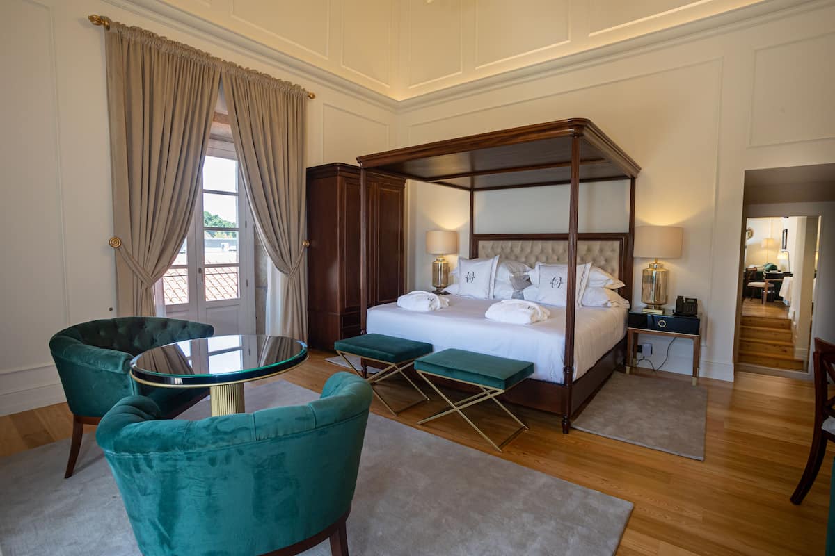 A spacious bedroom with a four-poster bed, a sitting area with a glass table, and a view through a large window.