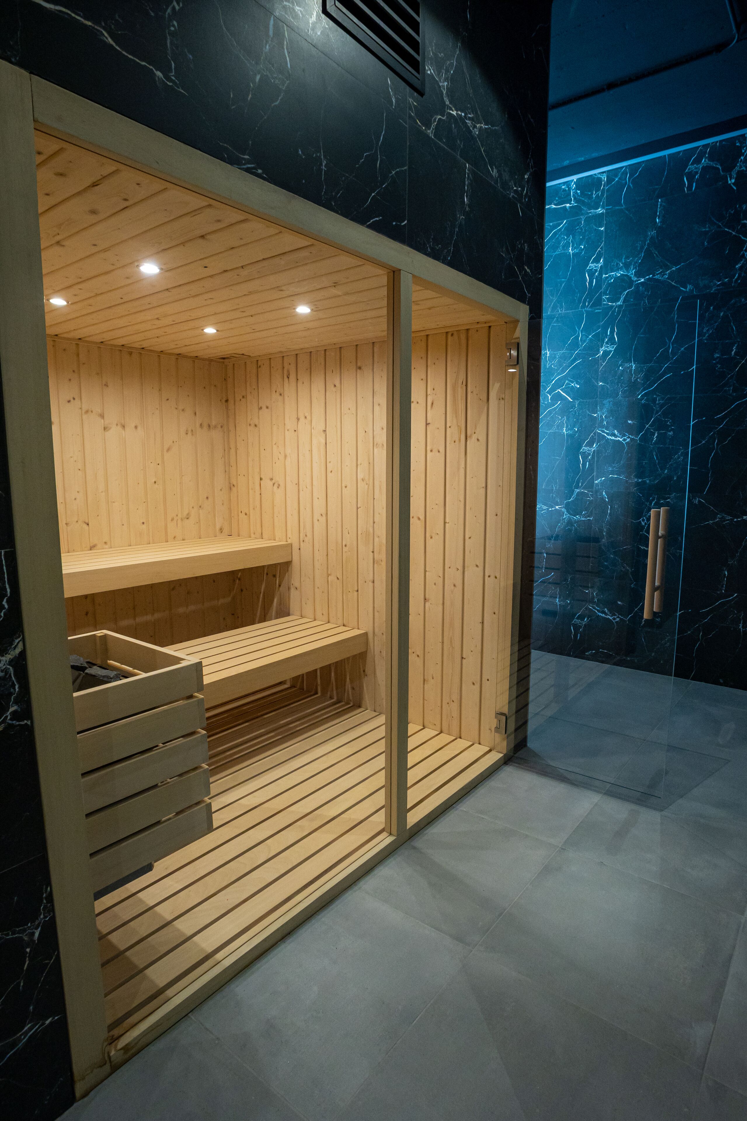 A sauna with wooden benches and a glass door, set against a dark marble wall.