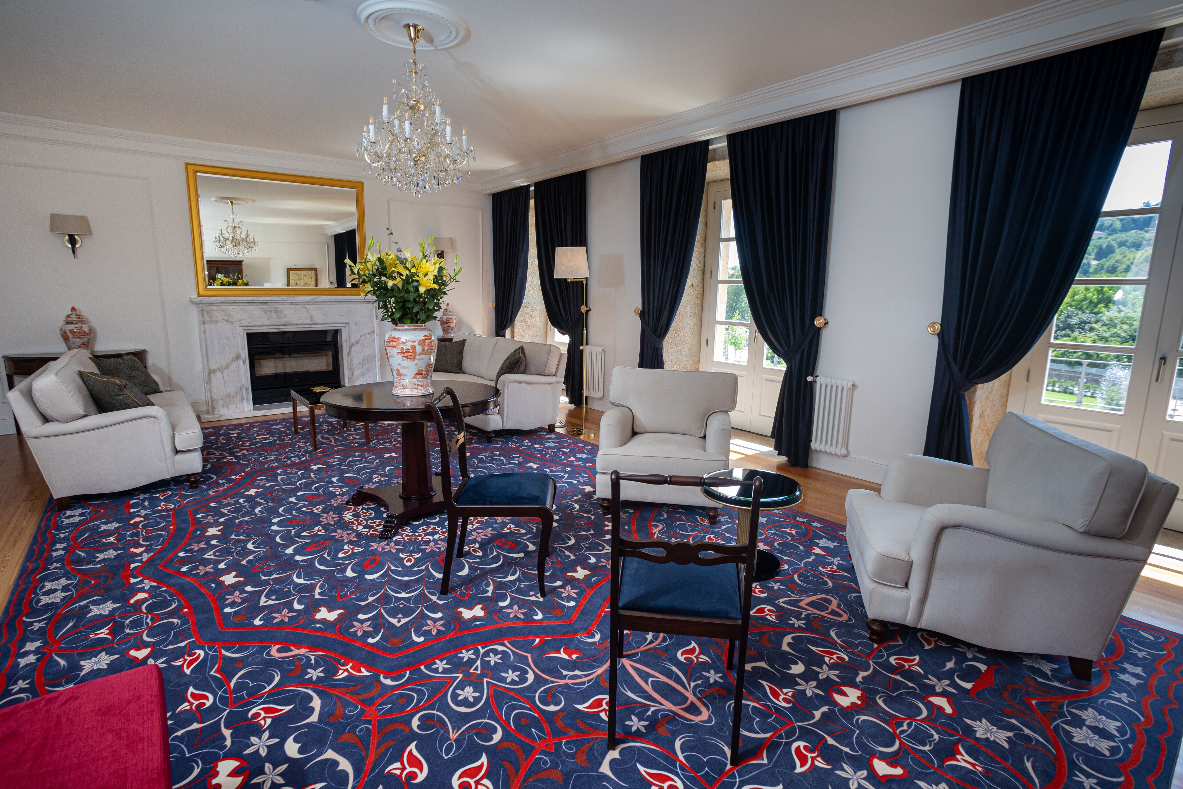 Un salón amplio con un tapete de diseño, diversas opciones de asiento, una chimenea y ventanas grandes con cortinas oscuras.