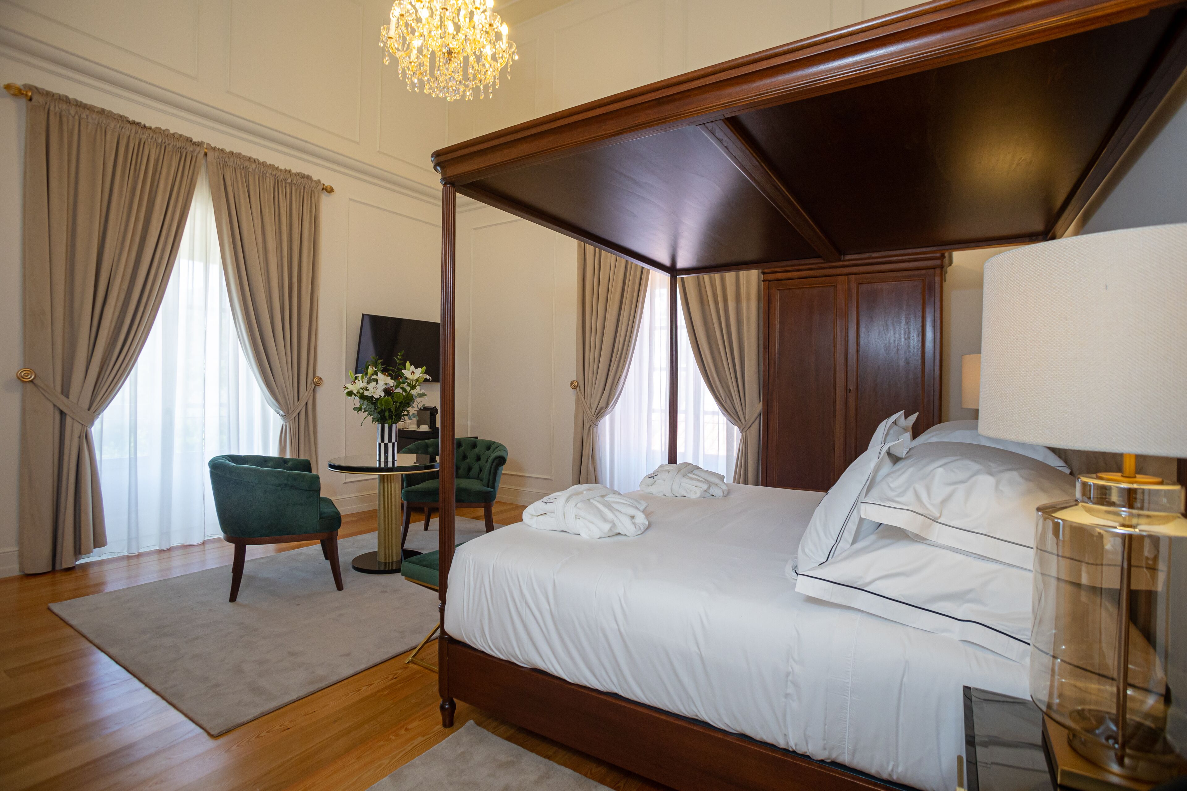 Una cama con dosel y sábanas blancas, un sillón verde, un escritorio con un florero y una araña.