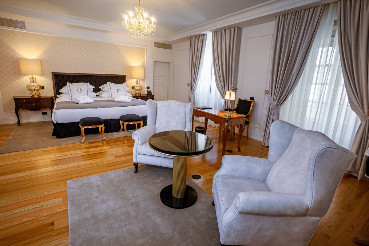 A hotel room with a bed, two armchairs, a round table, a desk, and a chandelier.