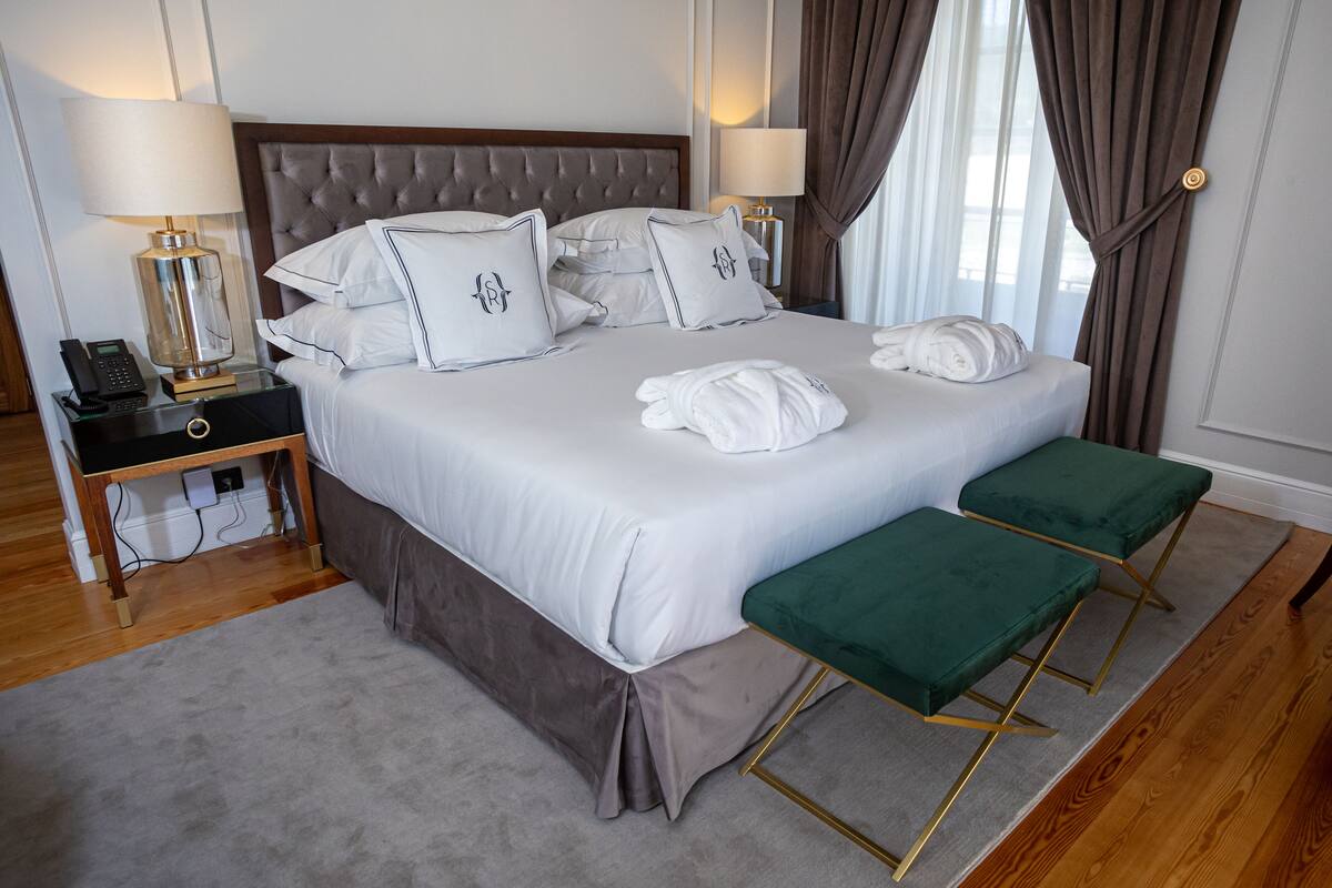 A neatly made bed with white linens and matching pillows, a bedside table with a lamp, a green bench, and a wooden floor.