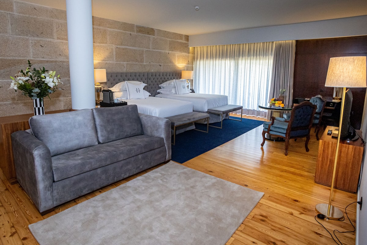 A hotel room with a grey sofa, a bed with white linens, a small table with a lamp, and a wooden floor.