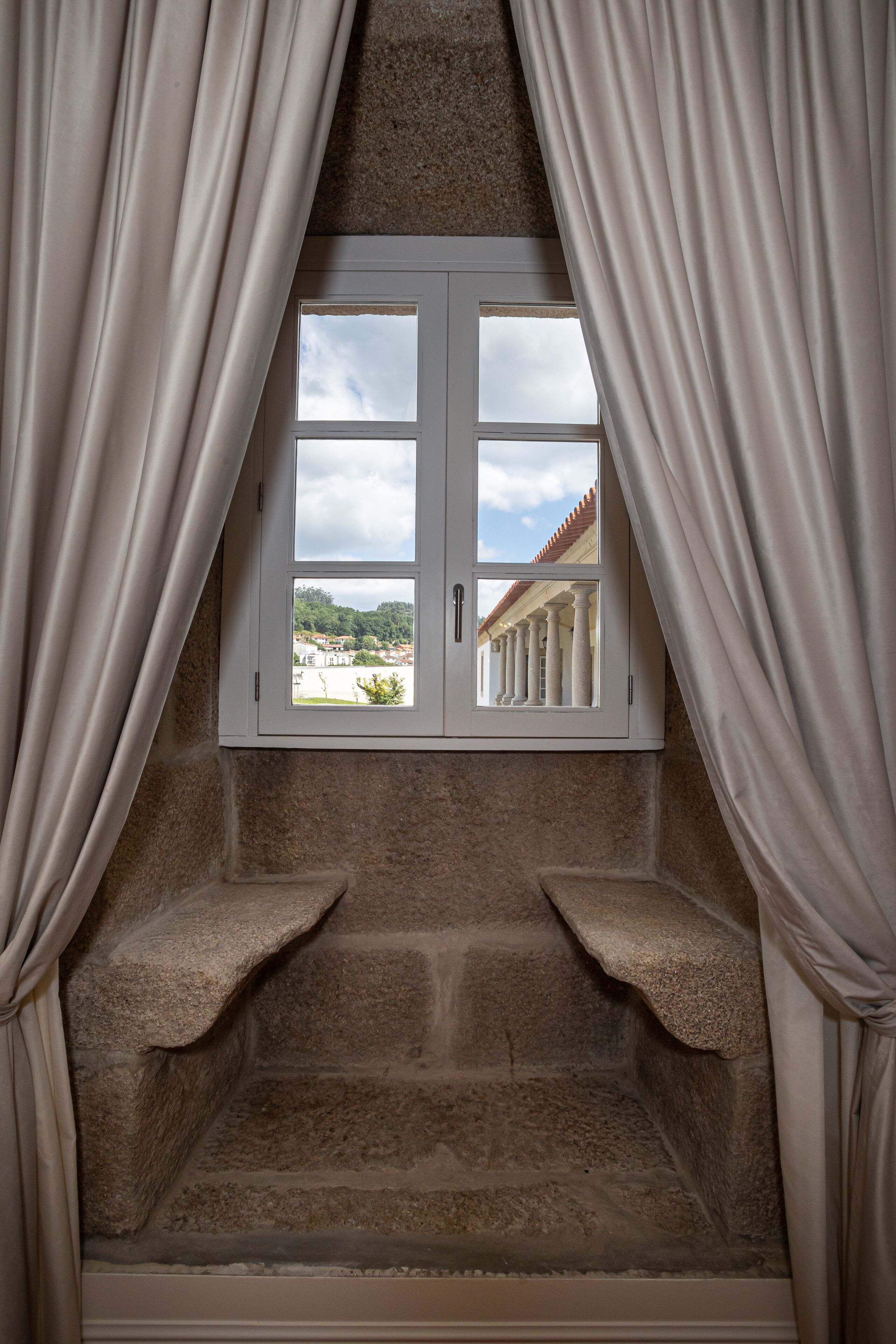 Um recanto com paredes de pedra, uma janela e cortinas, com vista para um edifício com colunas e um telhado de telhas vermelhas.