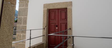 A stone building with a red door, a small balcony with a metal railing, and a clear blue sky in the background.
