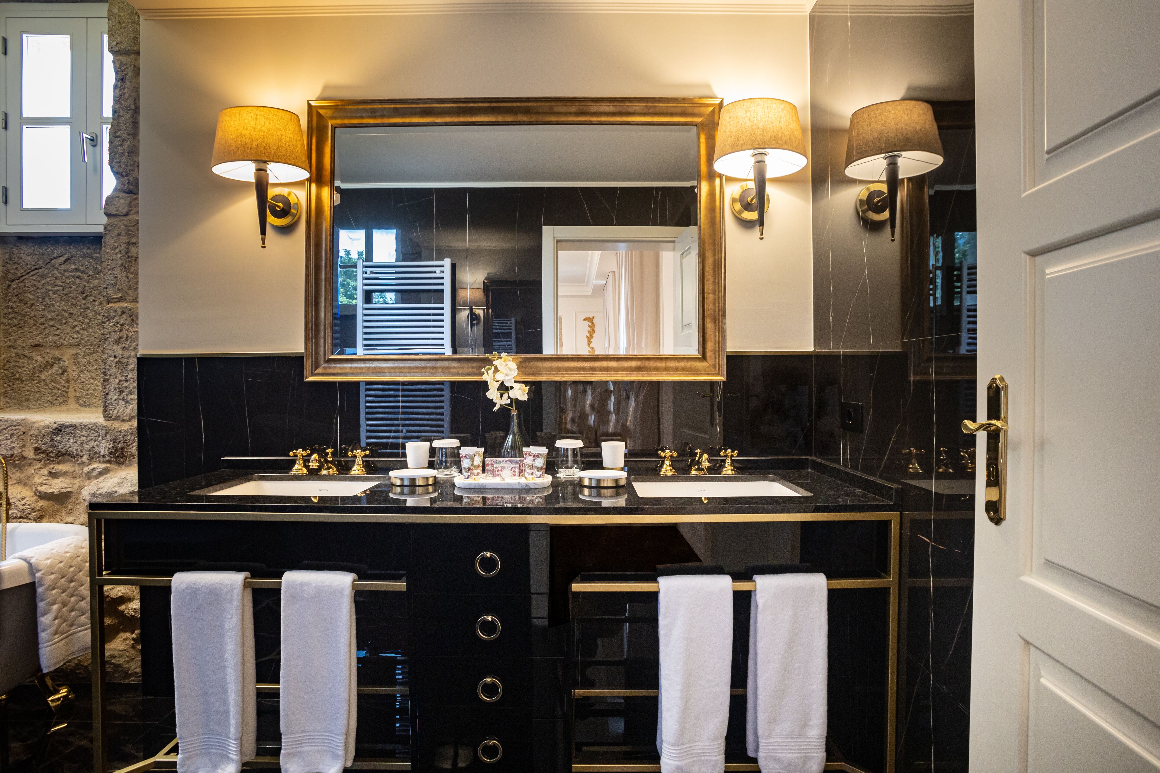 A bathroom with a black marble vanity, two sinks, and a large mirror with wall sconces. There are towels hanging on the vanity and a shower visible in the background.
