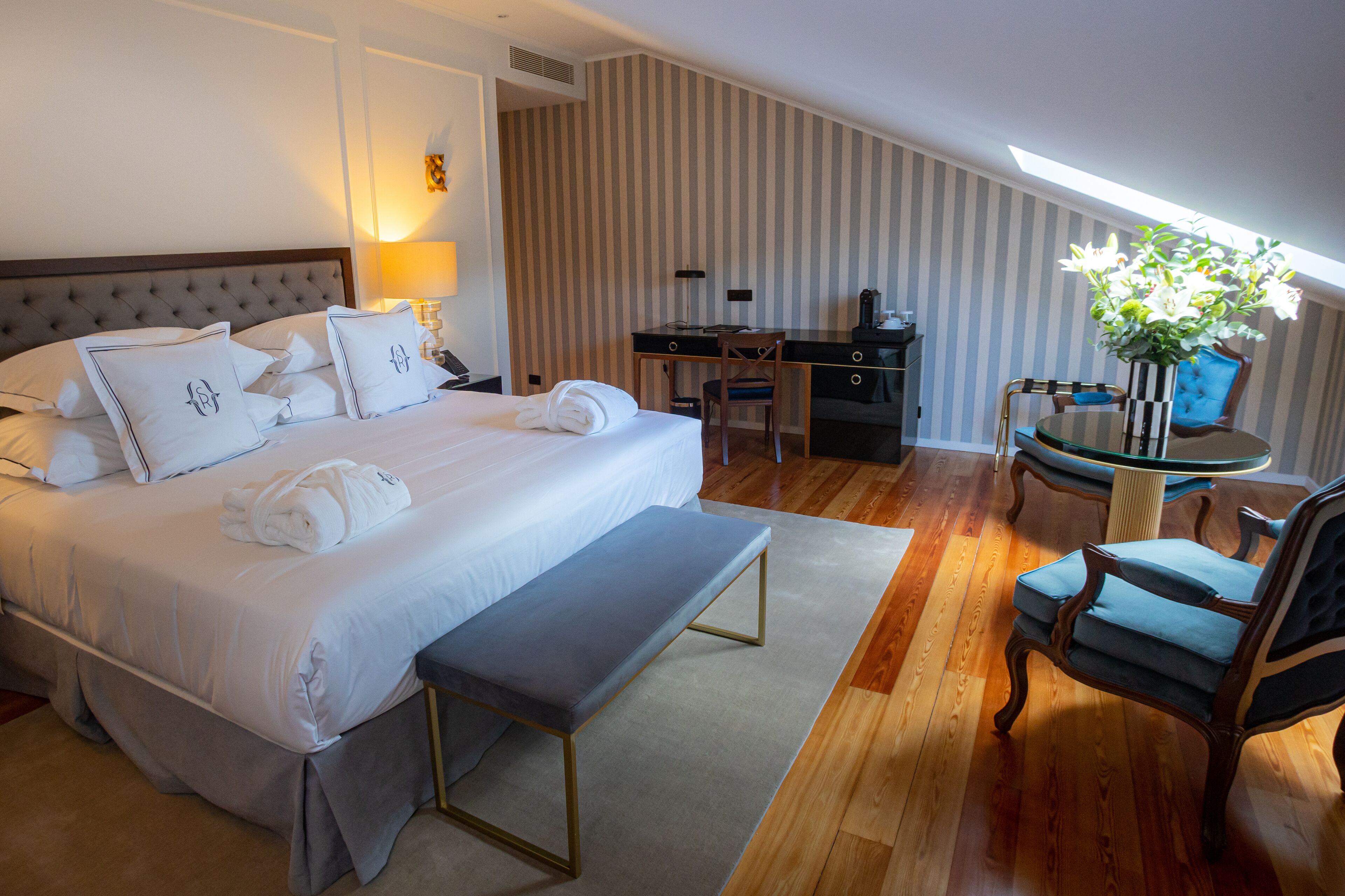 Hotelkamer met een groot bed, een bureau, een stoel, een bankje en een kleine tafel met een bloemenvaas.