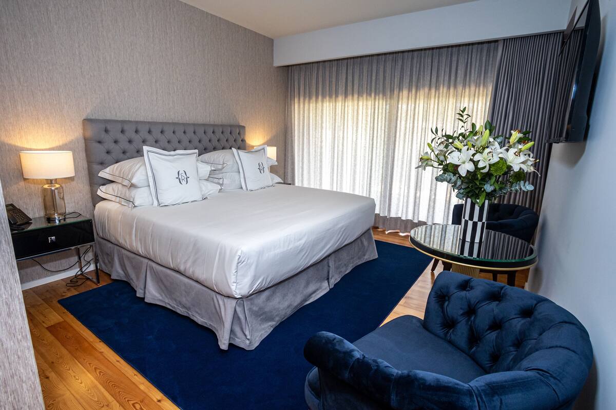 A hotel room with a large bed, a bedside table with a lamp, a round table, and a dark blue armchair.
