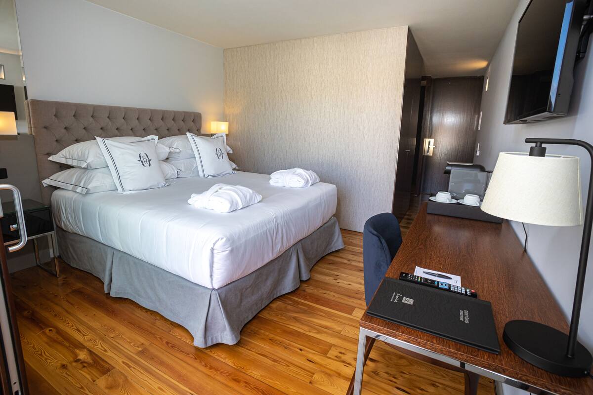A hotel room with a bed, bedside table, lamp, and a desk with a remote control.