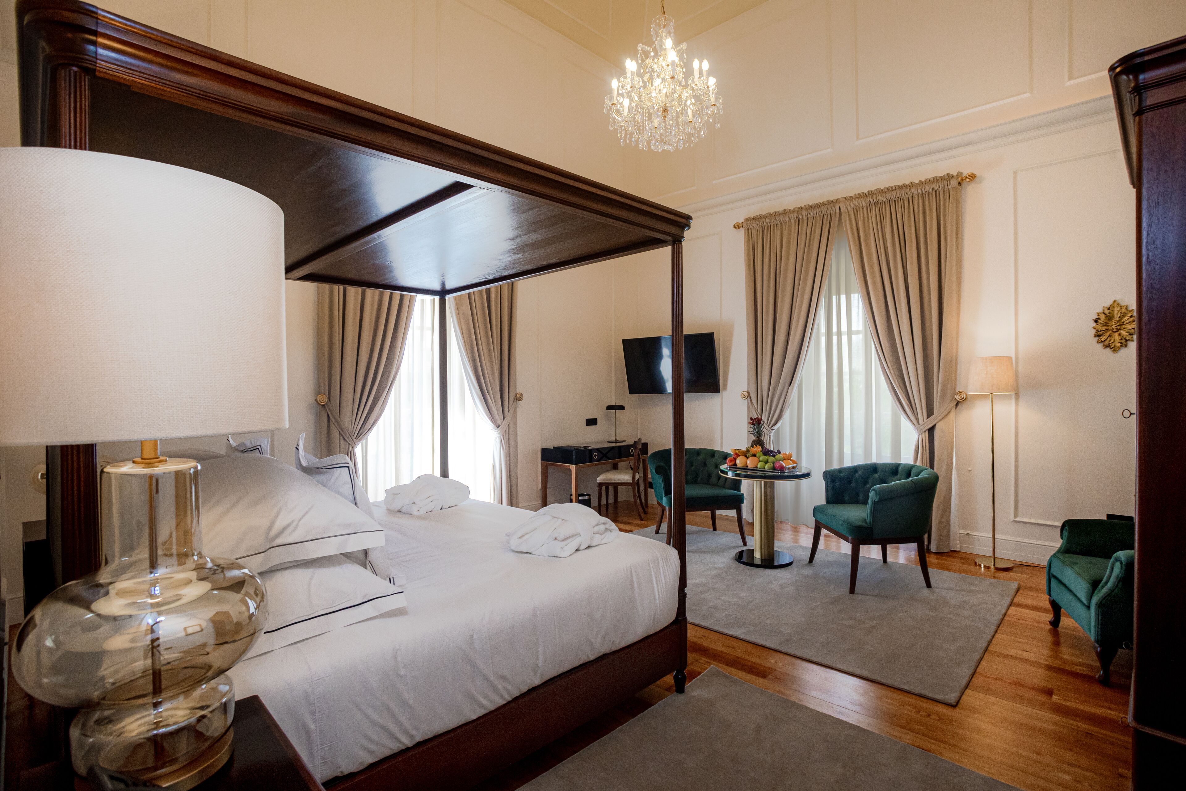Una cama con dosel, una lámpara de araña, un escritorio con televisor y dos sillones verdes en una habitación luminosa con suelo de madera.