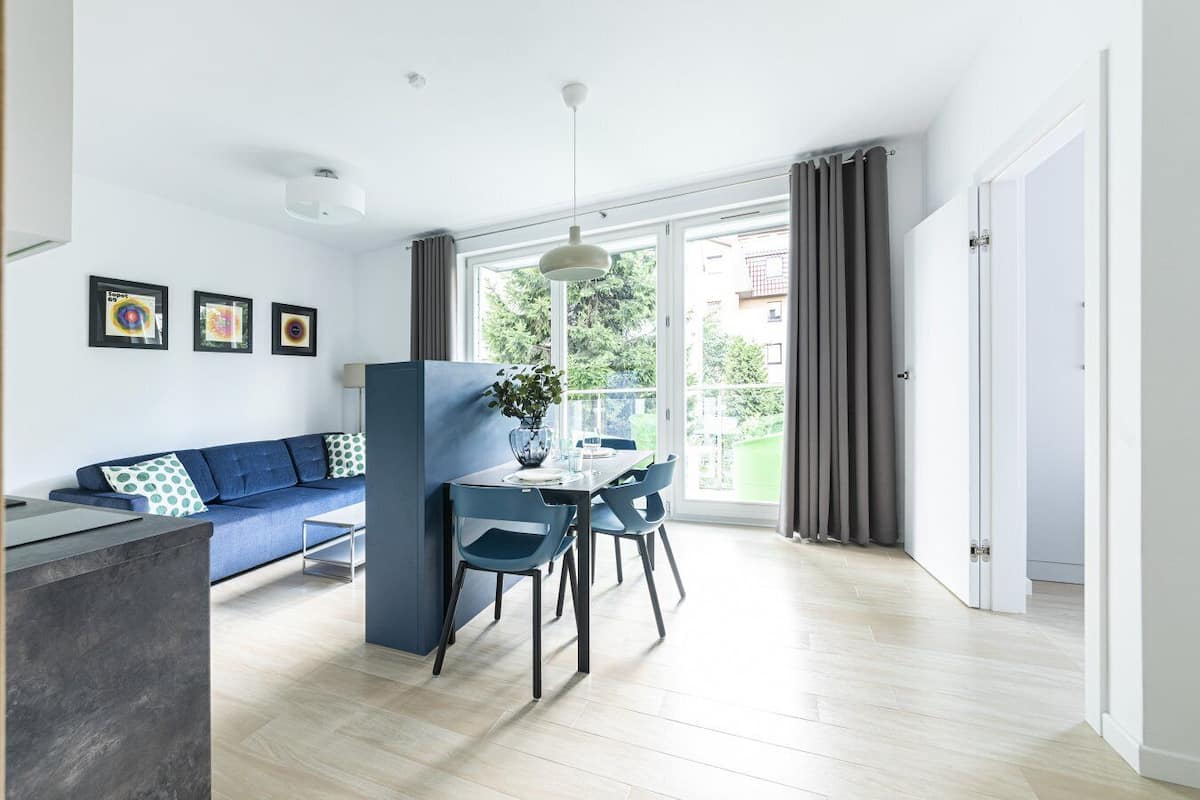 A modern living room with a blue sofa, a dining table with chairs, a pendant light, and a view of the outdoors through large windows.