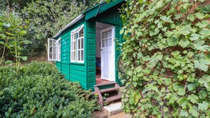 A small, green wooden house with a white door and windows, surrounded by lush greenery and a brick wall. - Warren Cottage (Bures)