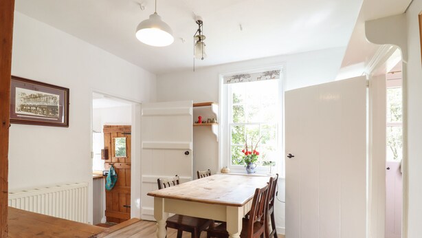 A dining area with a wooden table and chairs, a white radiator, a hanging light fixture, and a window with a view of greenery. - Warren Cottage (Bures)