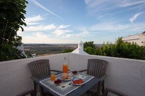 A table with breakfast items and a bottle of orange juice on a balcony overlooking a scenic landscape.