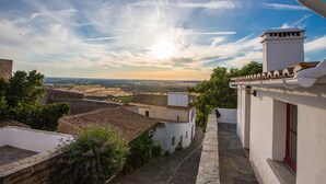 On aperçoit un bâtiment traditionnel, avec un chemin de pierre menant à une porte blanche, qui surplombe un paysage rural composé de champs en terrasses et d’un ciel dégagé.