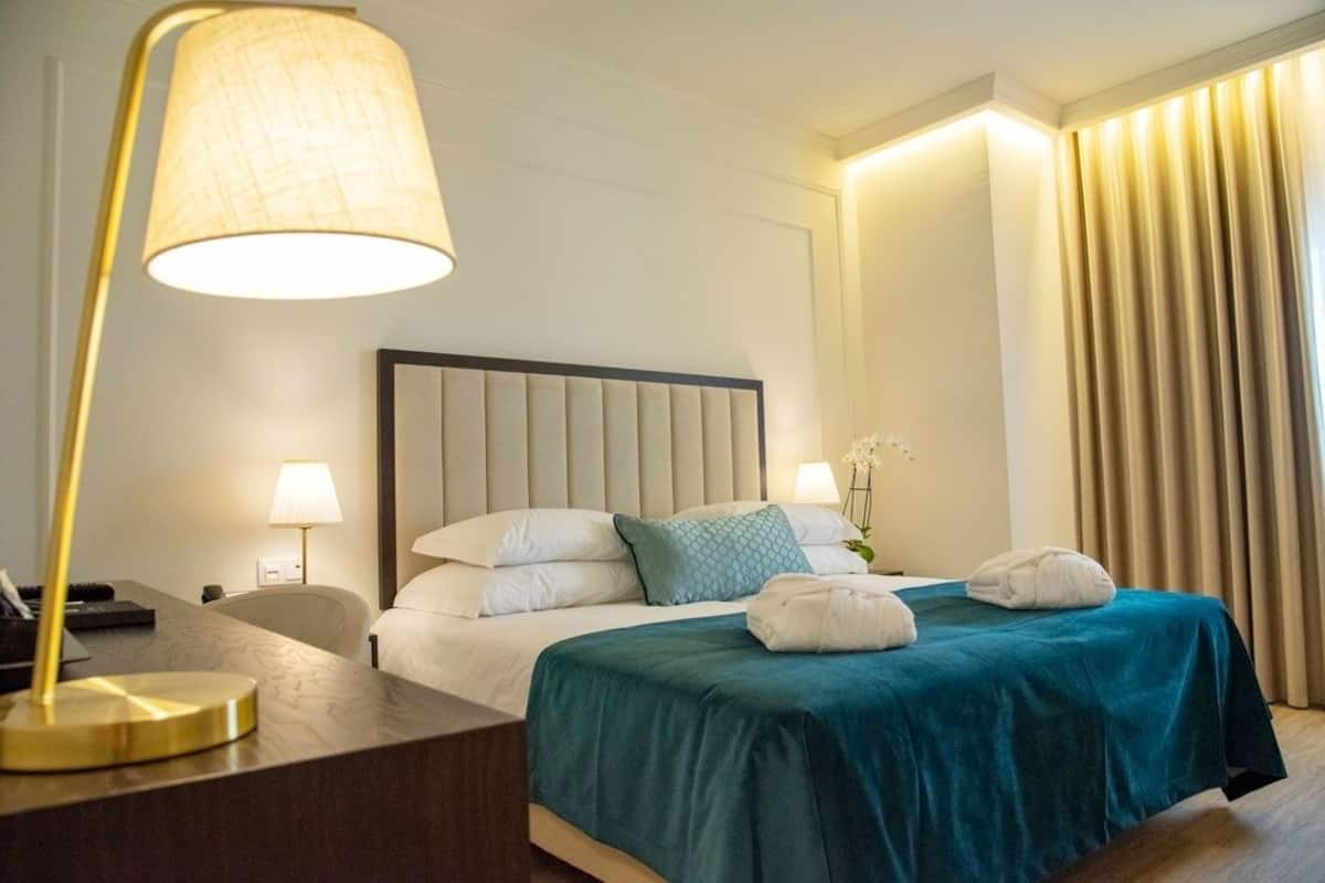 Quarto de hotel com uma cama grande, duas luminárias de cabeceira, uma escrivaninha com abajur e vista para as cortinas.