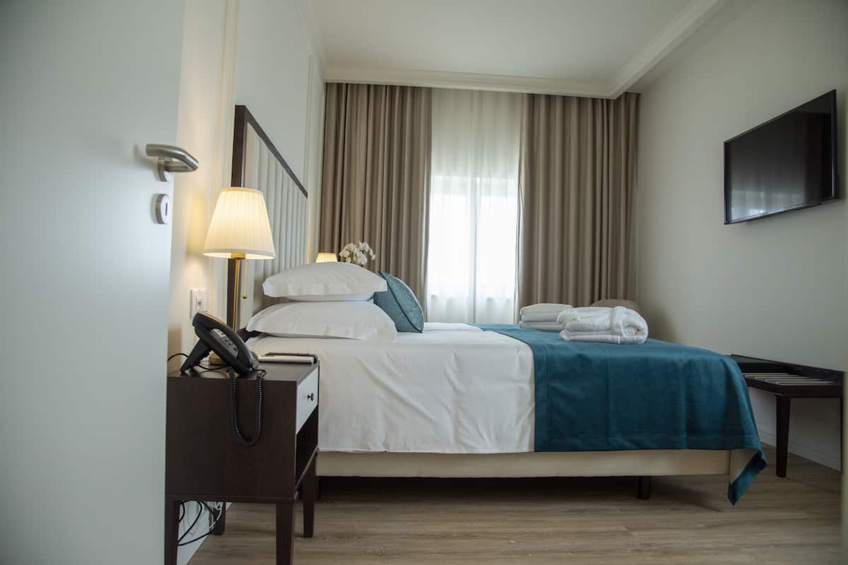 Quarto de hotel com cama, secretária com candeeiro, telefone e televisão na parede.