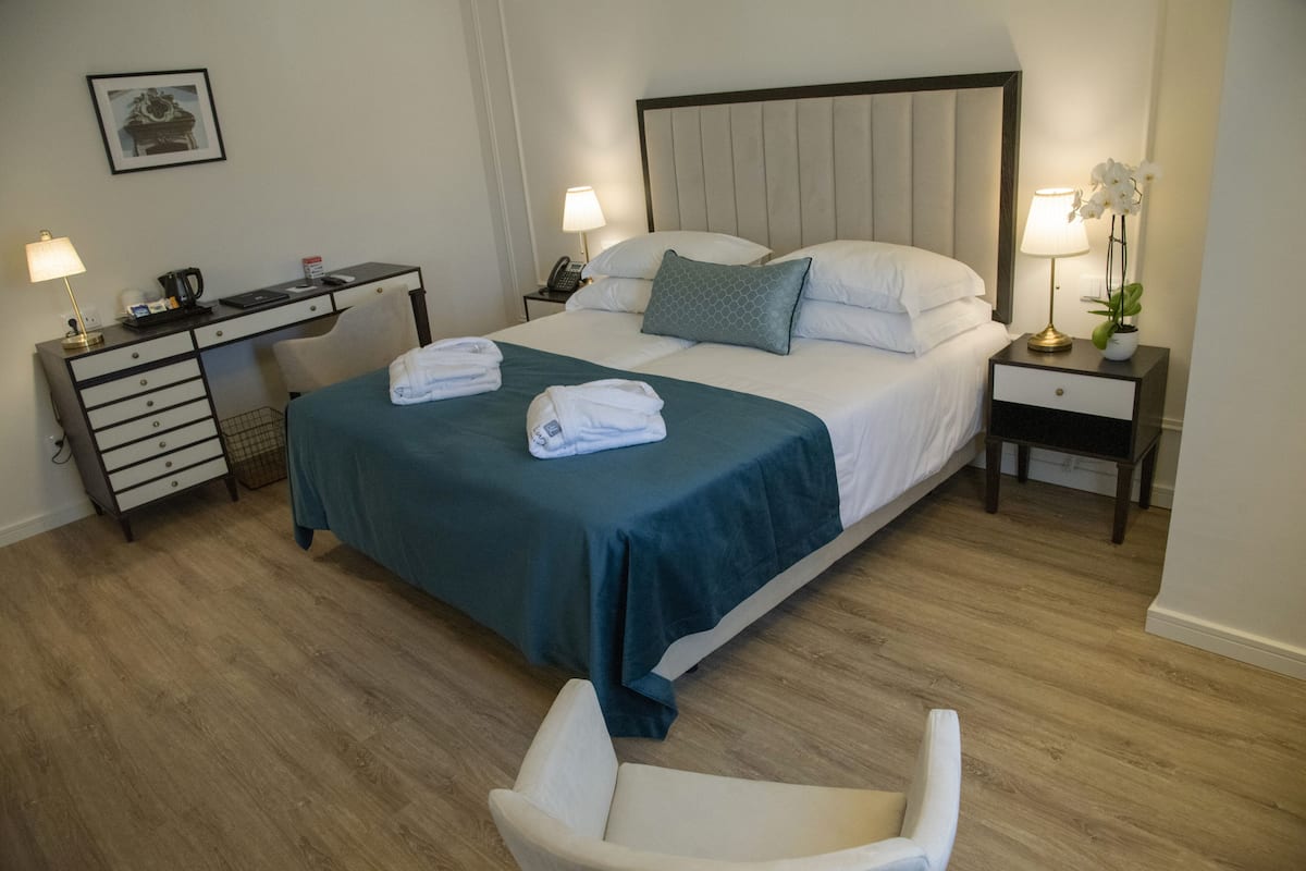 Quarto de hotel com uma cama, duas mesas de cabeceira, uma escrivaninha, uma cadeira e um abajur.