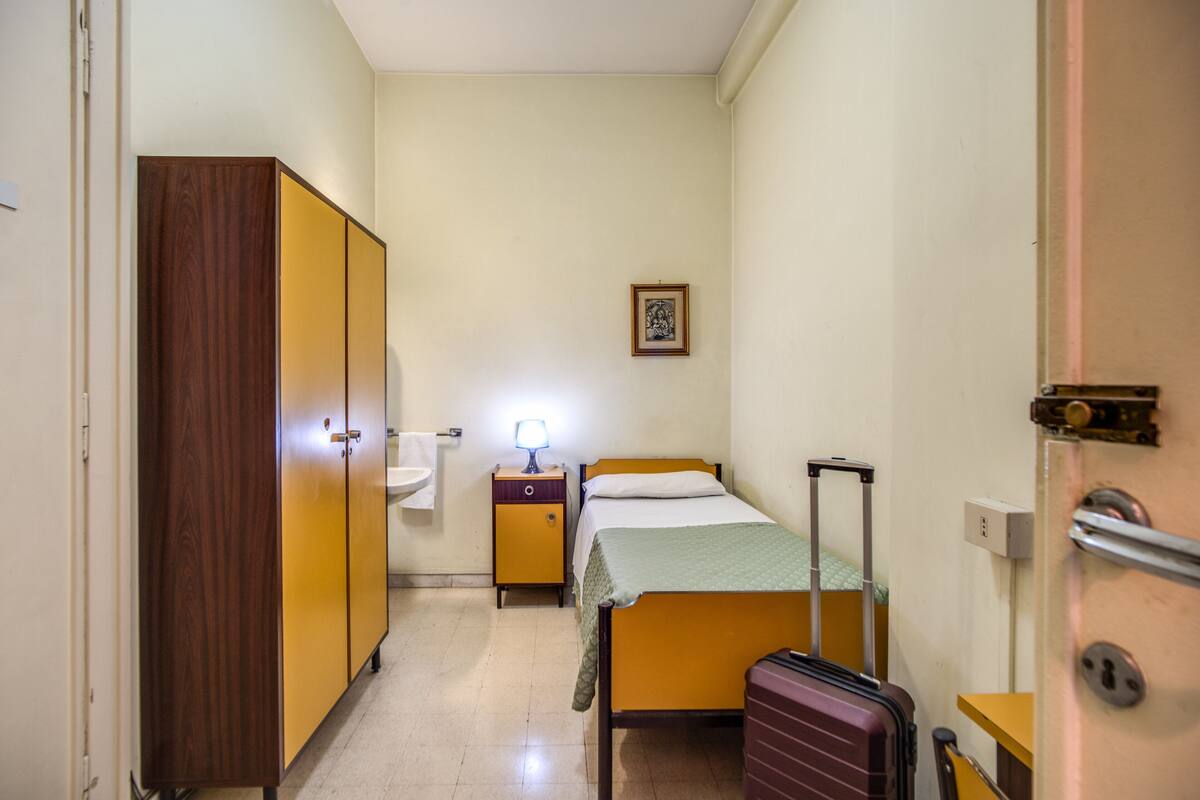 A small, narrow hotel room with a single bed, a wooden wardrobe, a nightstand with a lamp, and a suitcase.