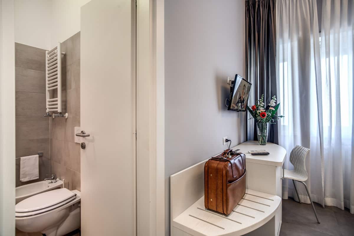 A bathroom with a toilet, a towel rack, and a shower area. There is a bench with a suitcase and a picture frame on it.