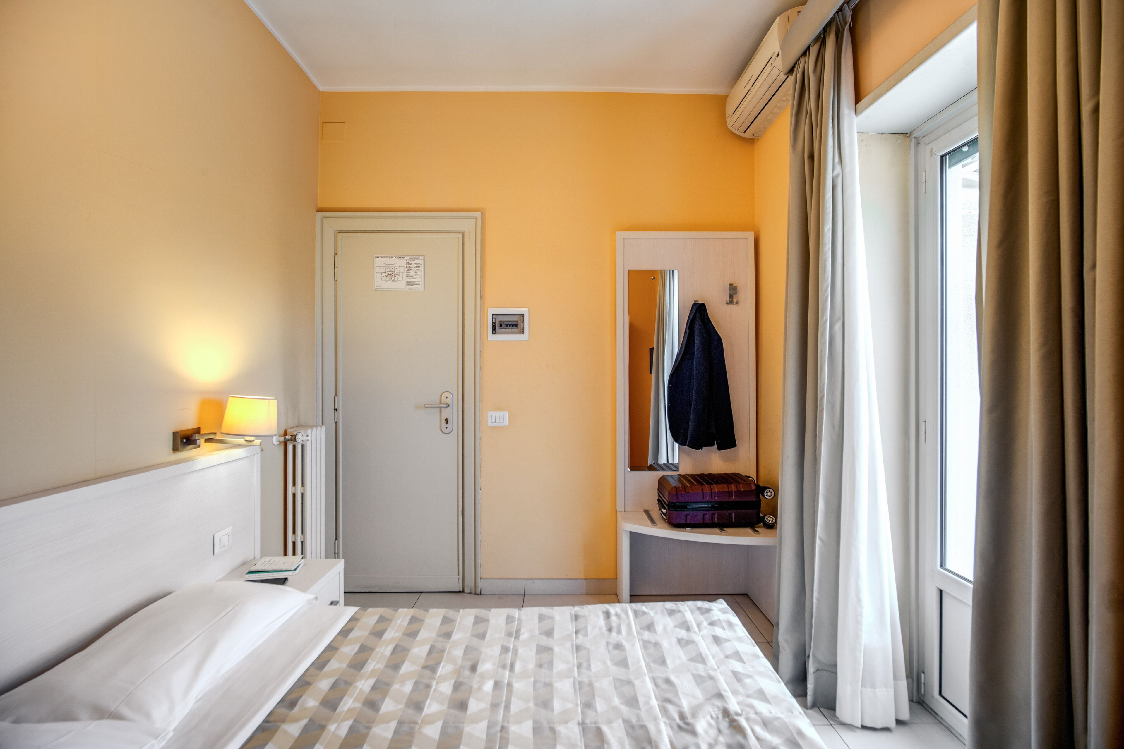 A hotel room with a bed, a nightstand with a lamp, a door, a mirror, a window with curtains, and a suitcase on a small table.