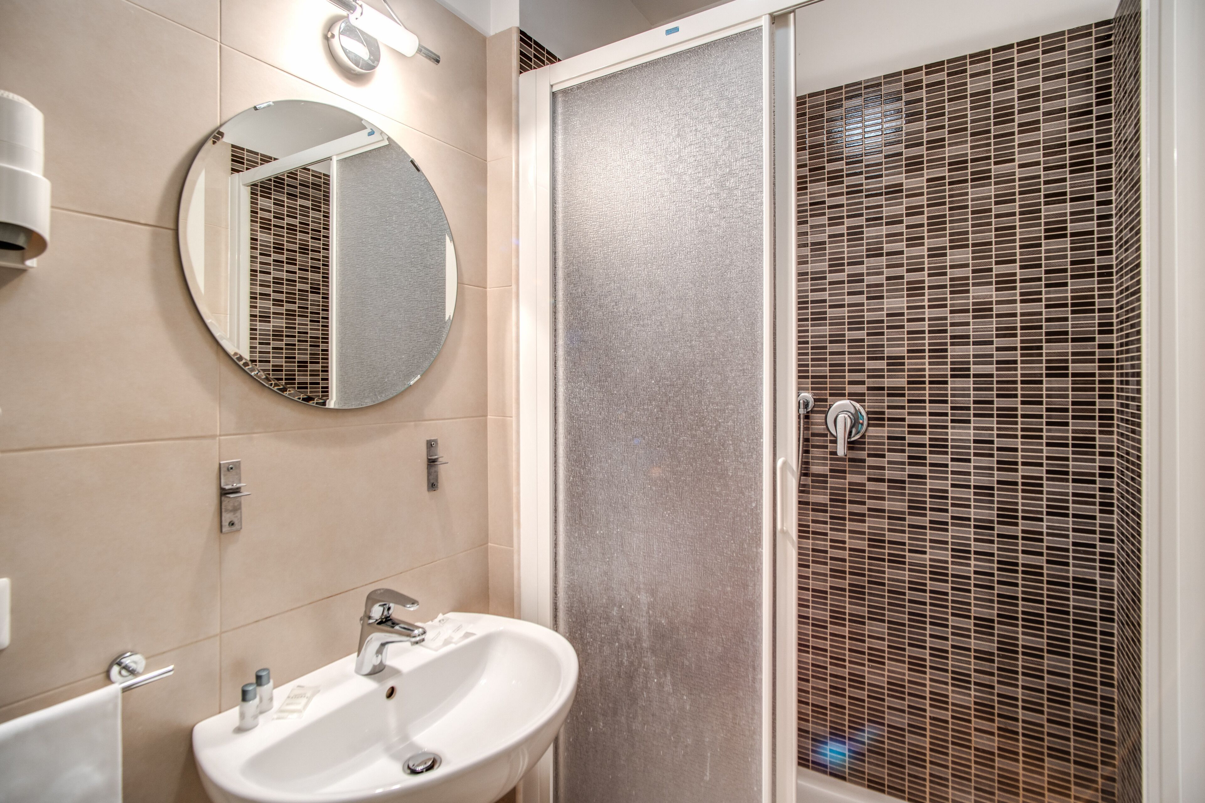 A bathroom with a round mirror, a sink, and a shower with a textured wall.